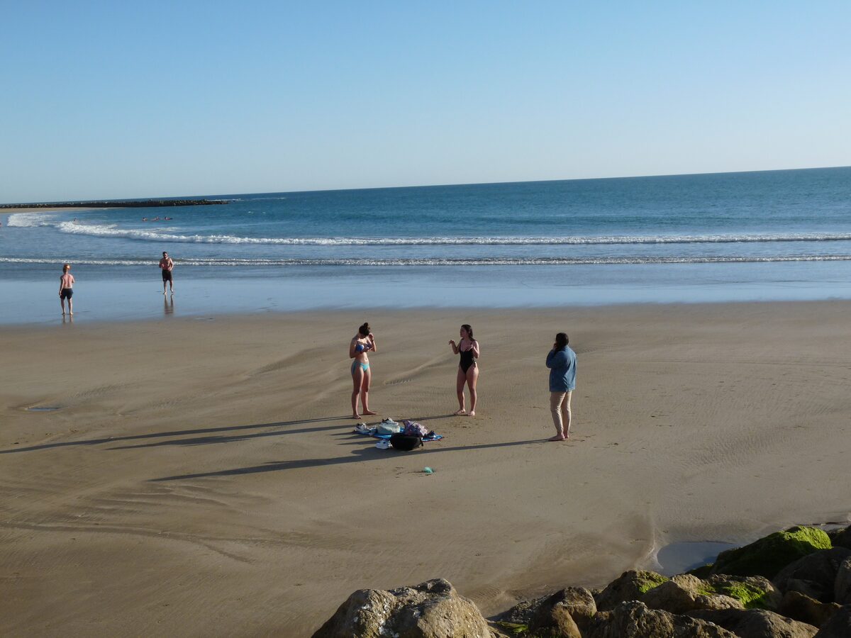 Sunny sand and small waves on the beach at Santa María del Mar, with a few people standing near the water and a breakwater behind