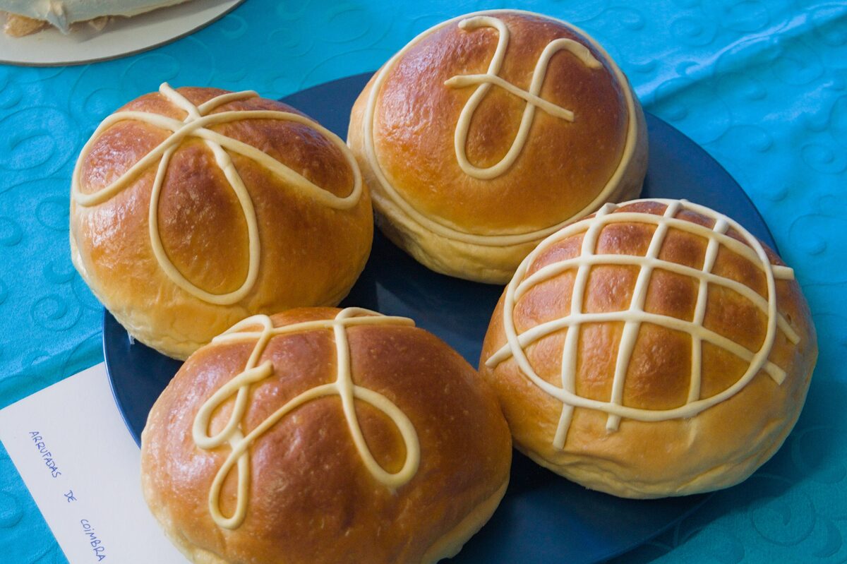 Four golden arrufadas sweet buns topped with bow and lattice patterns of white icing, on a dark plate over a turquoise tablecloth