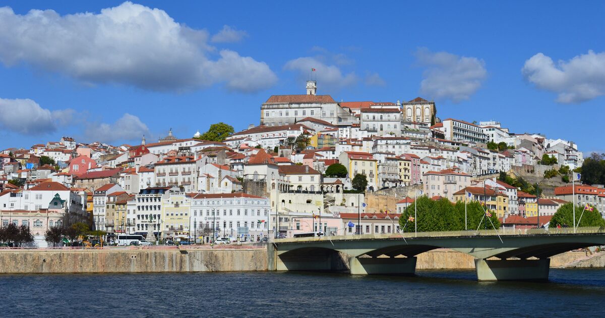 The old town of Coimbra piled up on its hill above the Mondego under a blue sky, with the university clock tower at the top and a road bridge in the foreground