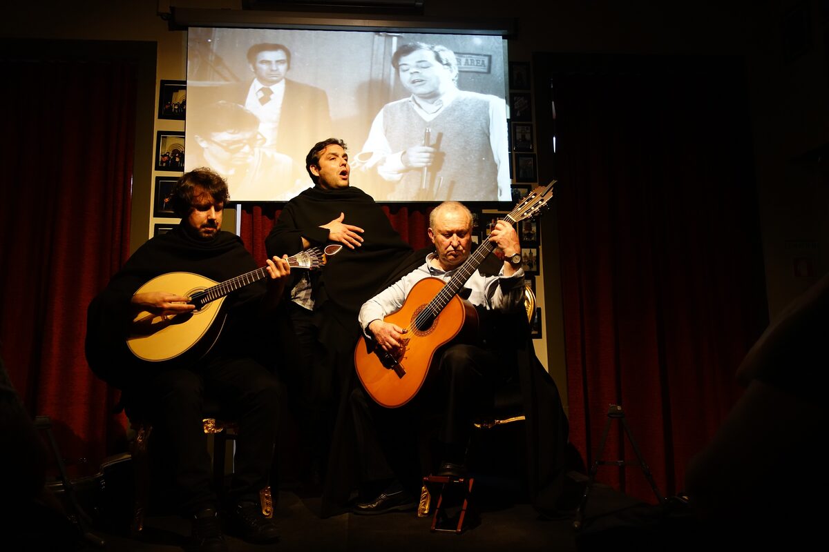 Three musicians in black student capes on a small stage, one playing the pear-shaped Coimbra guitar and one a classical guitar, with a screen showing black-and-white film footage behind them