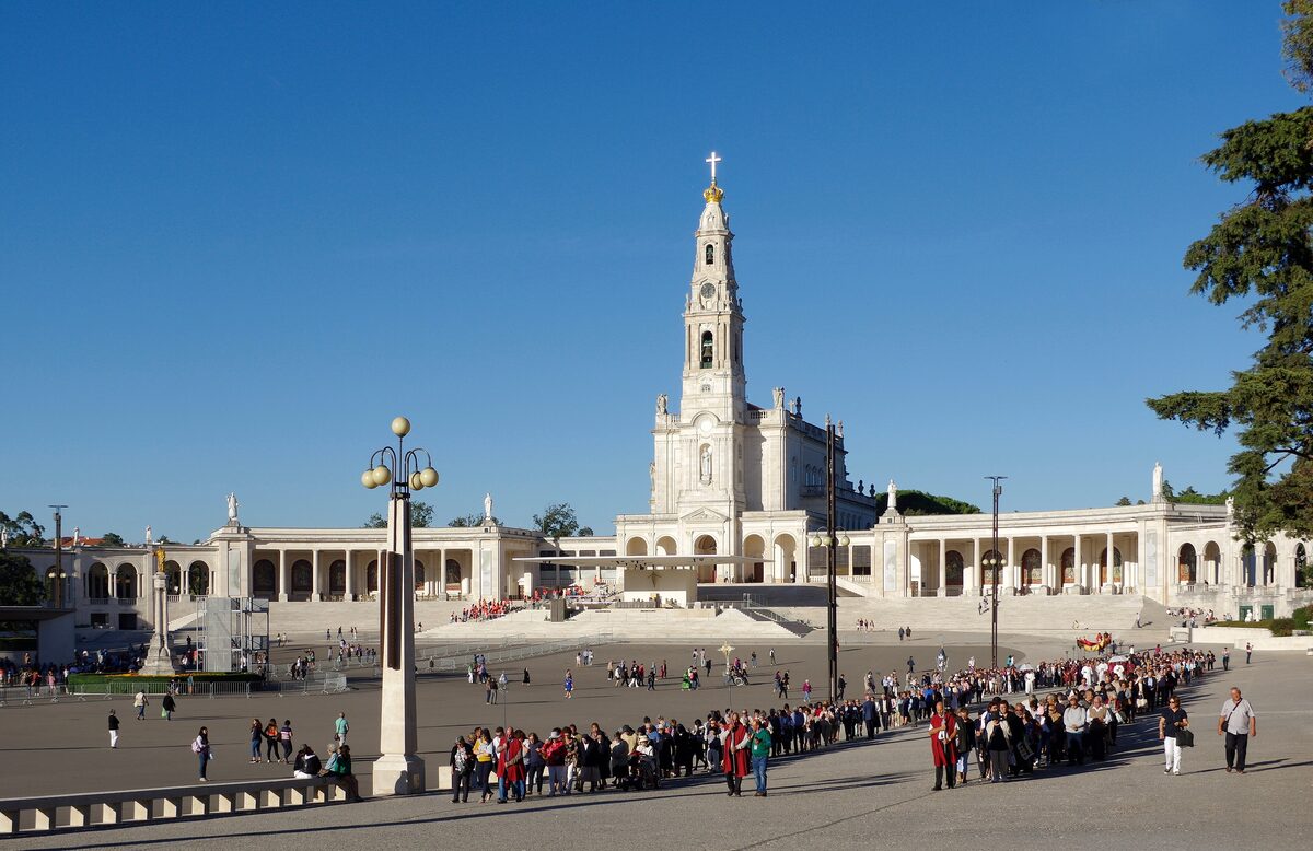 The white basilica with its tall tower and the curved colonnades of the Fátima sanctuary across the vast esplanade, with a line of pilgrims in front under a clear blue sky