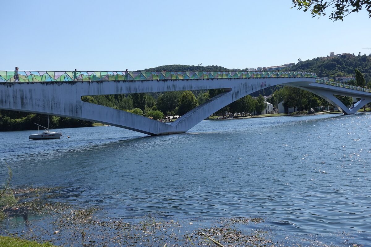 The Pedro e Inês footbridge in bright daylight, arching in two long spans over the blue Mondego, with a colored glass railing and a wooded hill behind