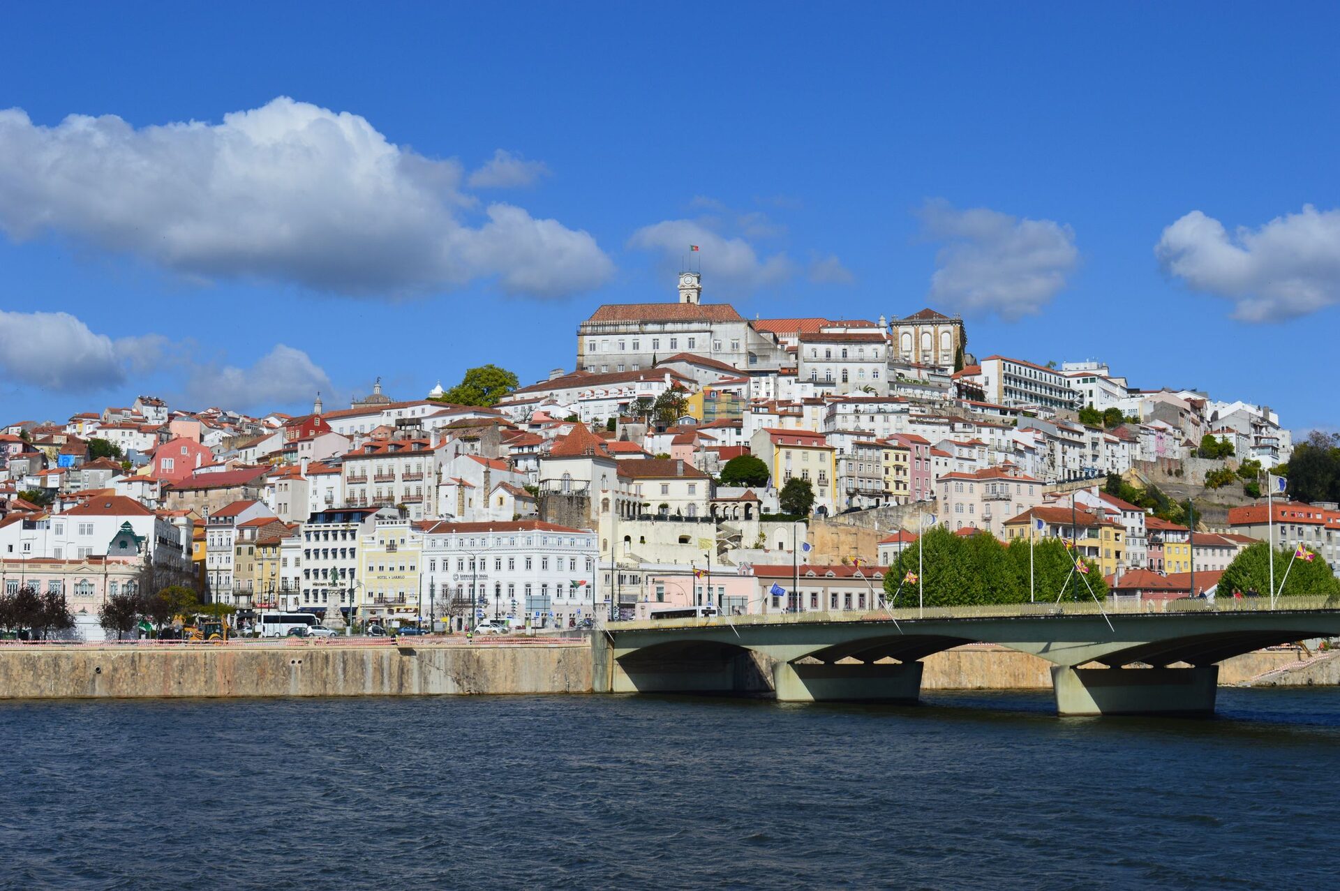 The old town of Coimbra piled up on its hill above the Mondego under a blue sky, with the university clock tower at the top and a road bridge in the foreground