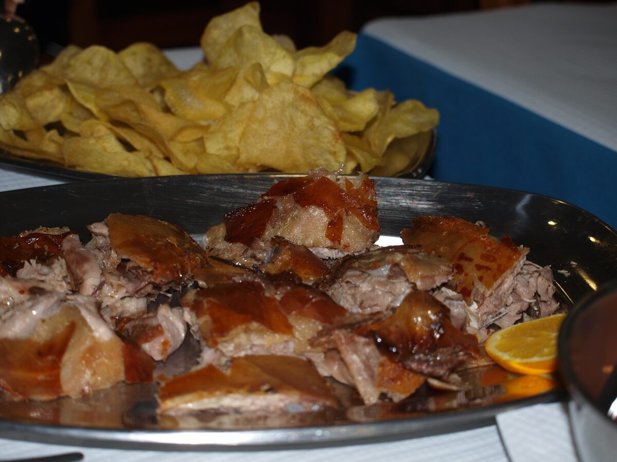 Chopped roast suckling pig on a metal platter, with glossy golden crackling skin on the pieces and an orange slice at the edge, with a dish of potato chips behind