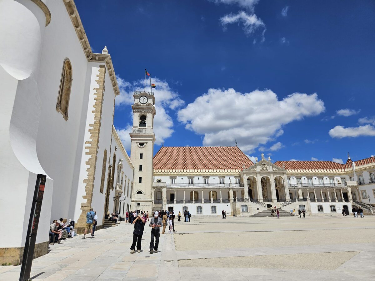 The courtyard of the old university palace under a blue sky, with the white wall of the chapel on the left, the clock tower and the red-roofed palace facade beyond, and visitors crossing the square