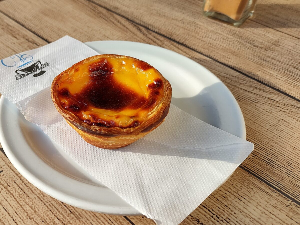 A single pastel de nata with a blistered, caramelized top on a small white plate and napkin, on a wooden cafe table beside a cinnamon shaker