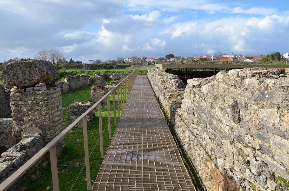 A metal walkway runs between the excavated Roman walls of Conímbriga, with grassy ground and the village beyond