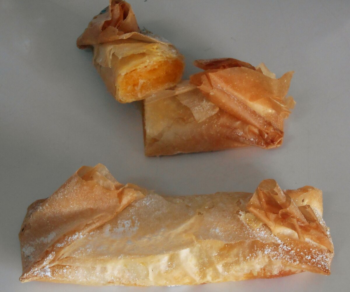 Three Tentúgal pastries on a grey surface, rolls of paper-thin pastry dusted with sugar, one cut open to show the orange egg-yolk filling