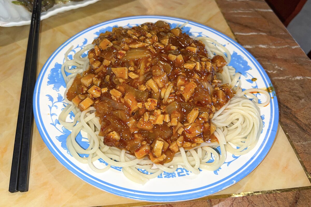 A plate of golden yellow noodles under a diced mushroom and meat sauce at a noodle restaurant in Dunhuang