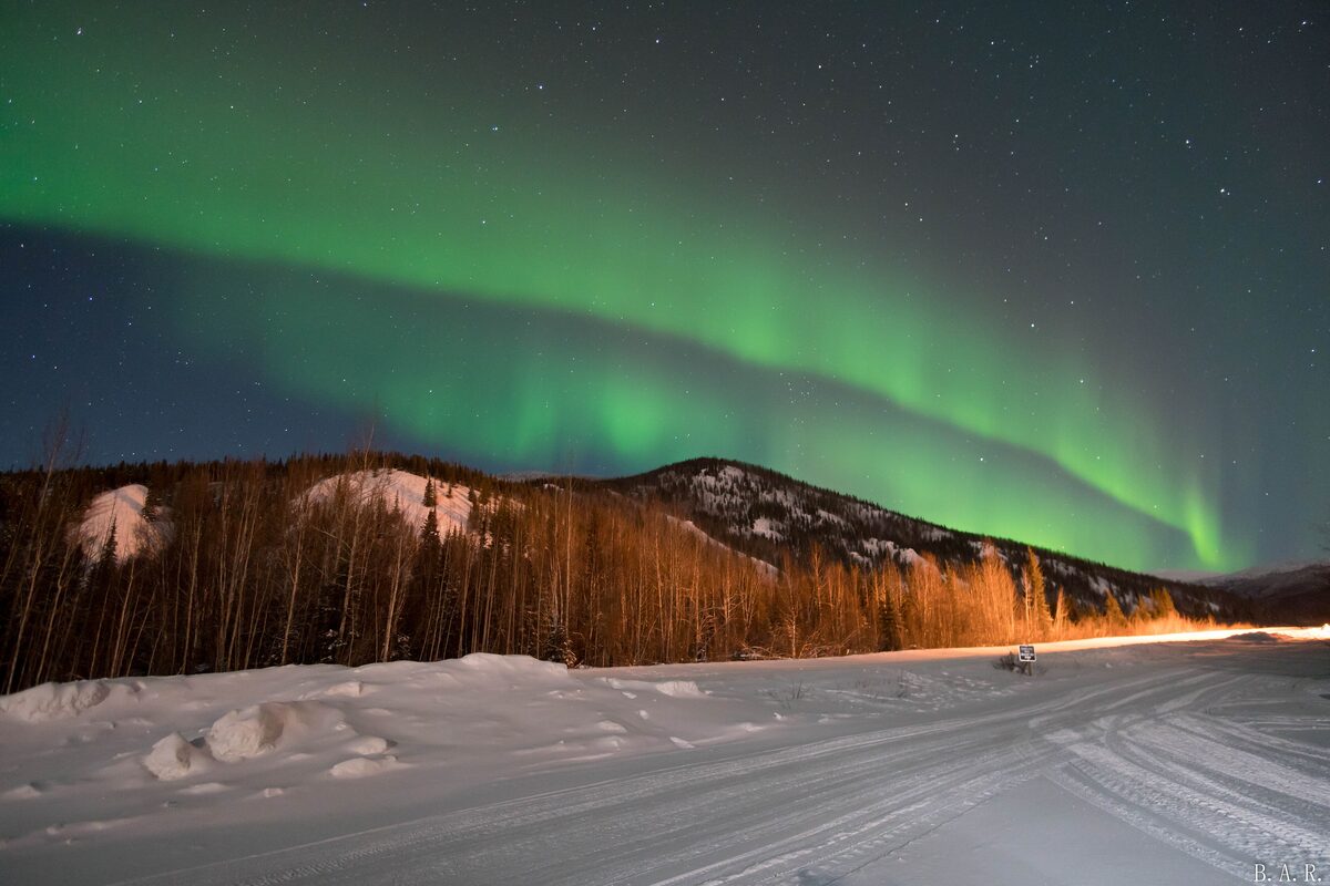 A green aurora arcing over snowy hills and a plowed track in the Chena Hot Springs area