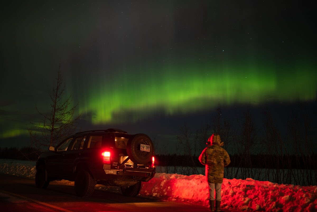 A person standing by a parked SUV on a snowy roadside, watching a green aurora arc across the night sky in Alaska