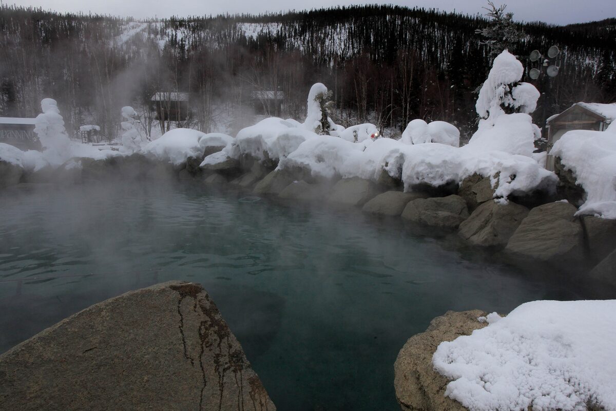 Steam rising from the rock-edged outdoor hot spring pool at Chena Hot Springs, with snow on the boulders and spruce forest behind