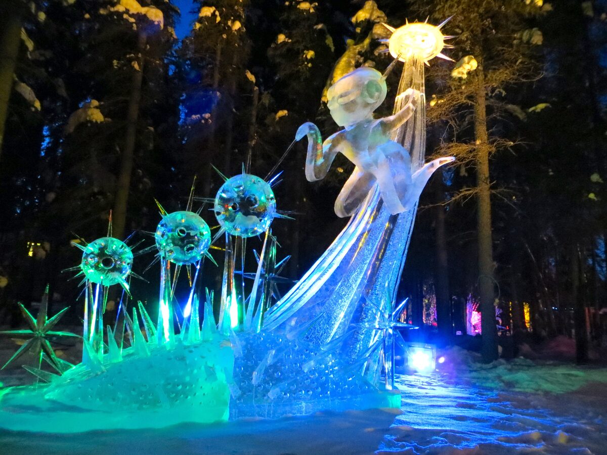 A lit ice sculpture glowing blue and green among spruce trees at night in Fairbanks