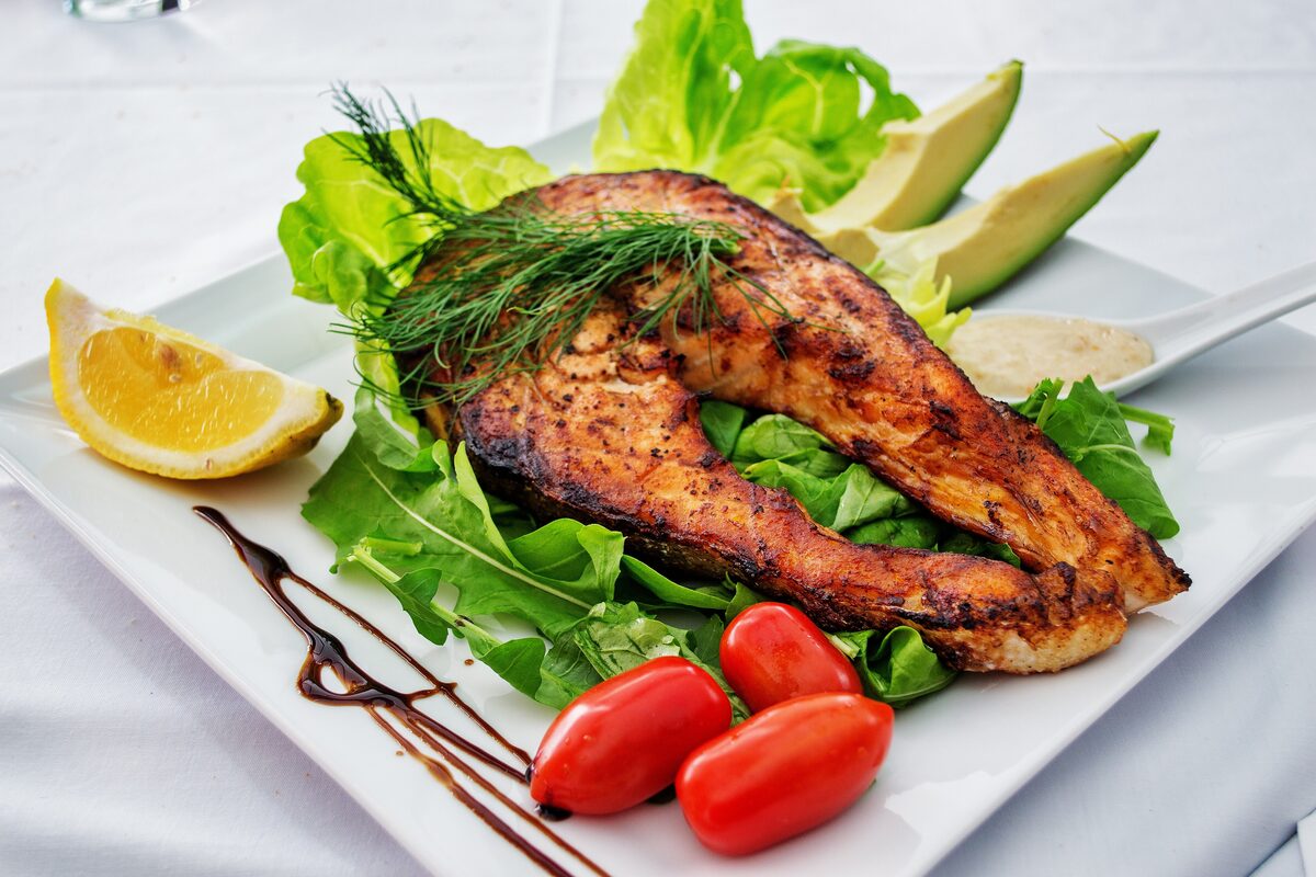 A grilled salmon steak on a plate with salad greens, avocado, tomatoes and a lemon wedge