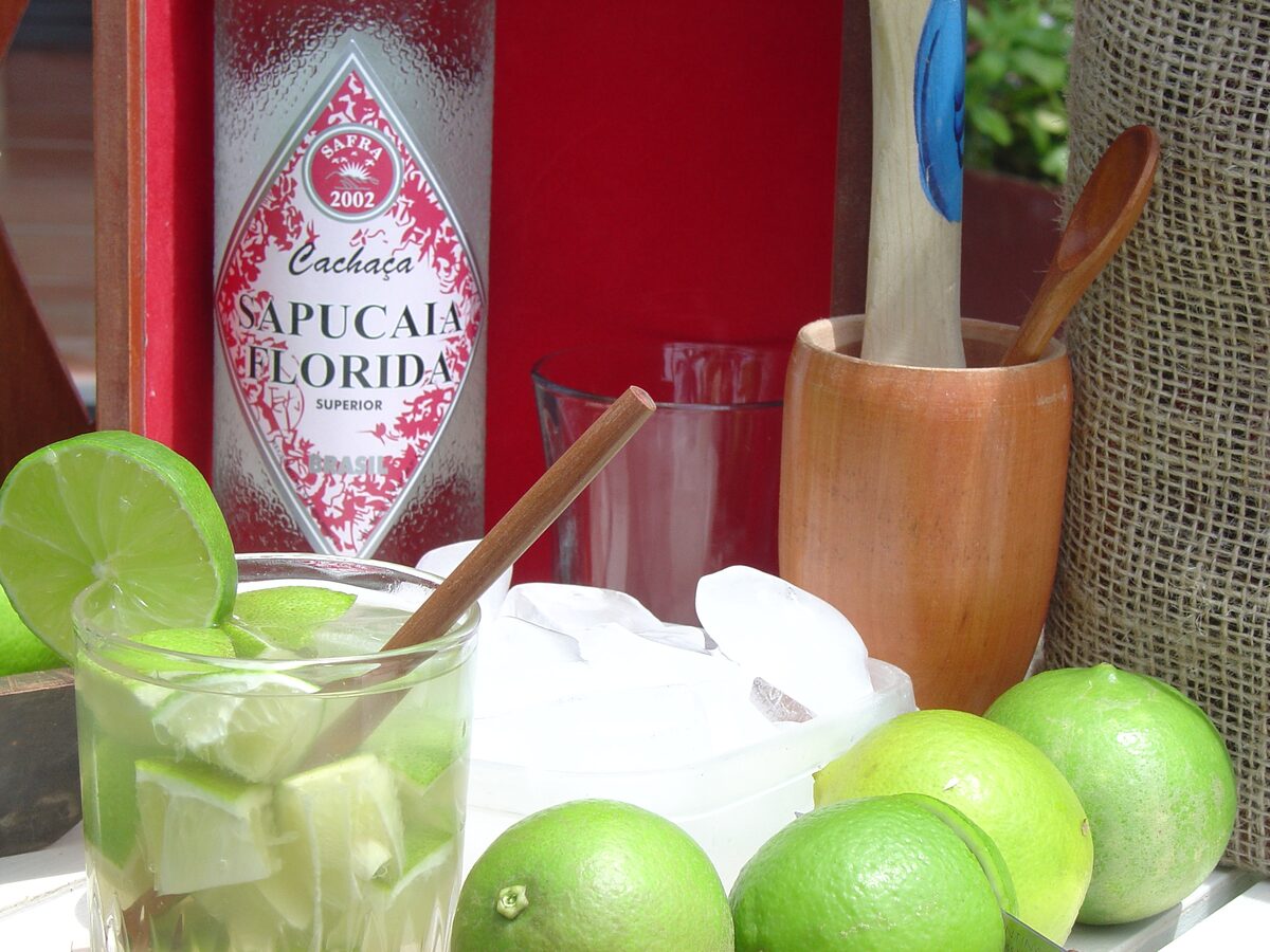 A caipirinha in a glass with lime wedges, a bottle of cachaça and a wooden muddler beside it