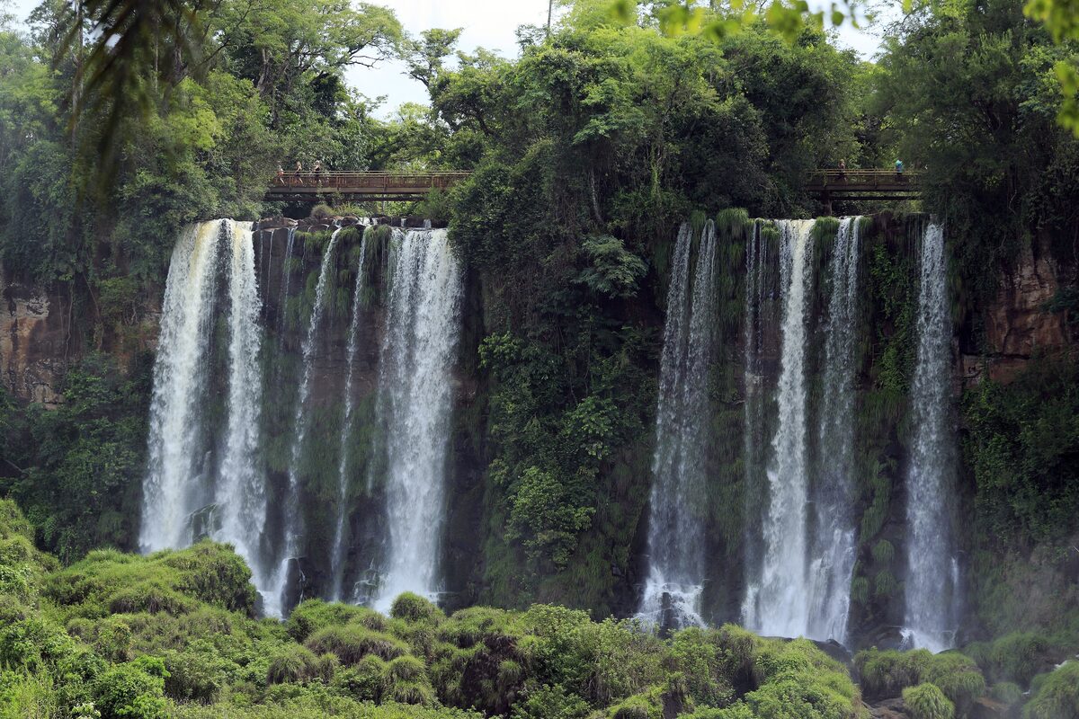 Two curtains of falls drop over a green cliff, seen from below on the lower circuit, with a small walkway on the cliff top