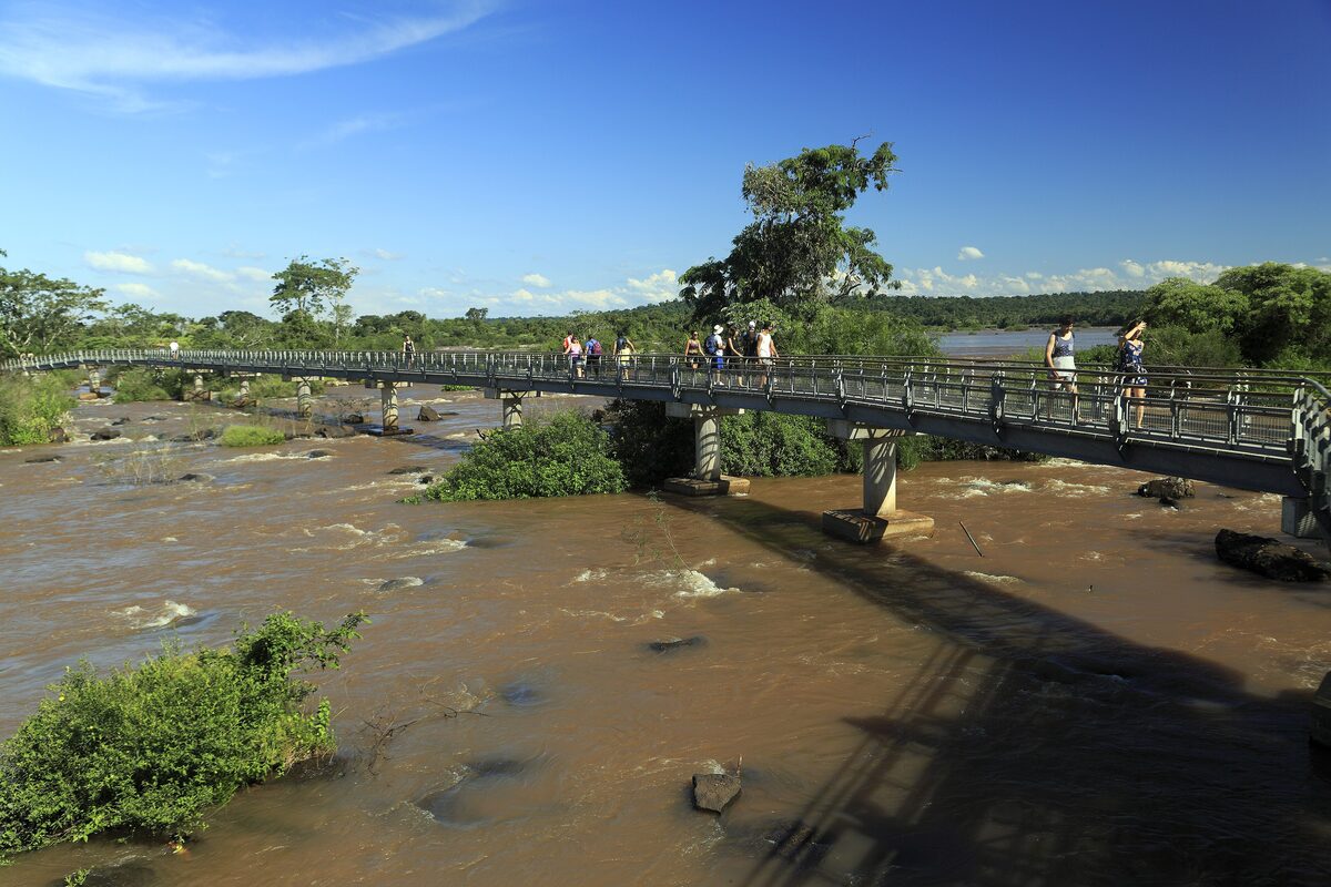 The upper circuit walkway crosses the broad, brown Iguazú river above the falls on the Argentine side, with visitors on the bridge