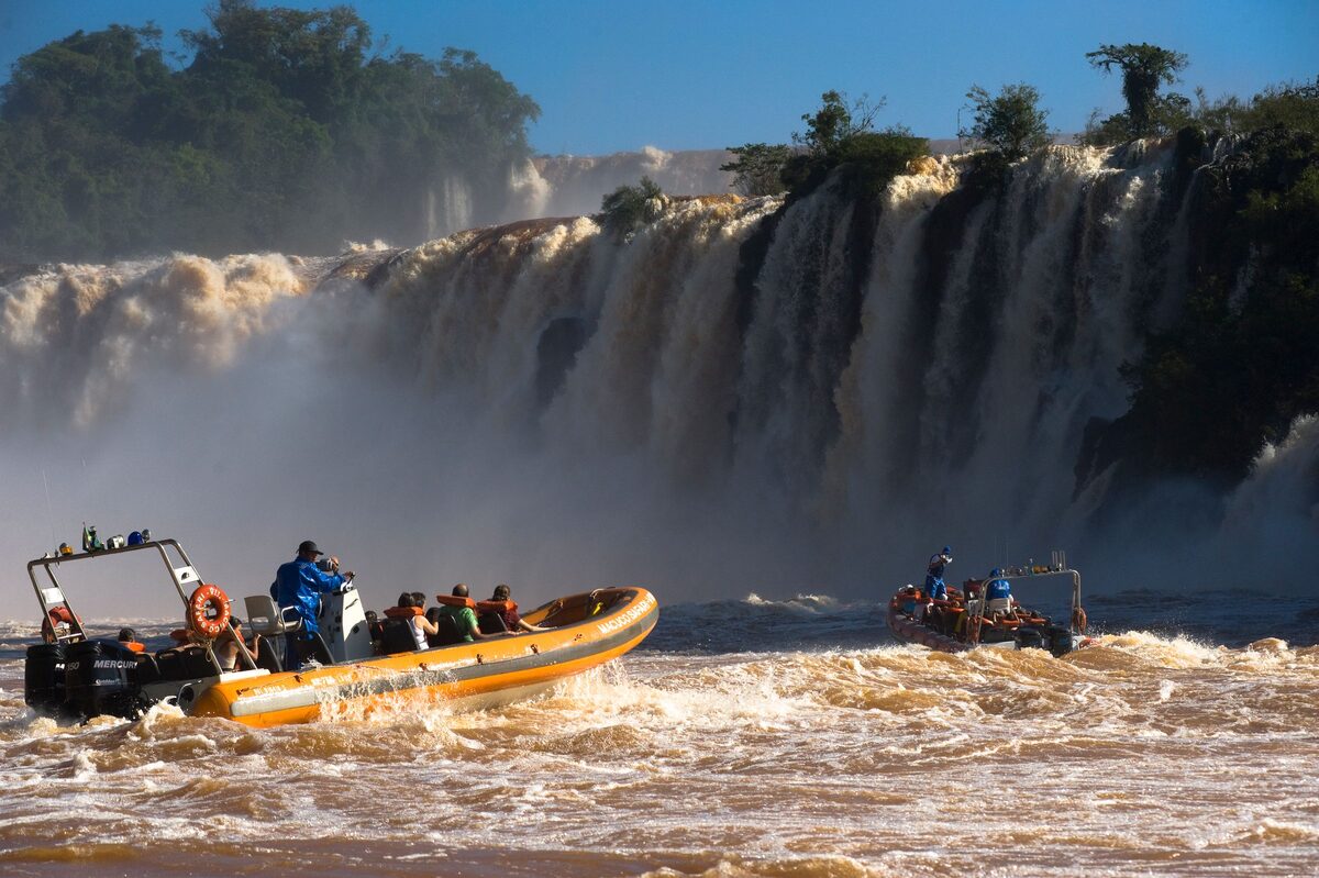 A motorized inflatable boat of tourists in life jackets at the foot of the curtain of falls, with a second boat behind