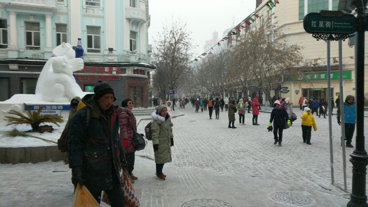 Walkers in winter coats on snowy Central Street, with a snow polar bear on the left and old European-style facades along the cobbled pedestrian street