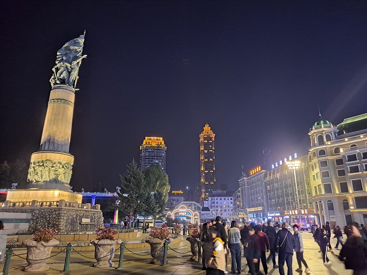 The lit Flood Control Monument column at night with a crowd on the plaza and lit high-rises behind