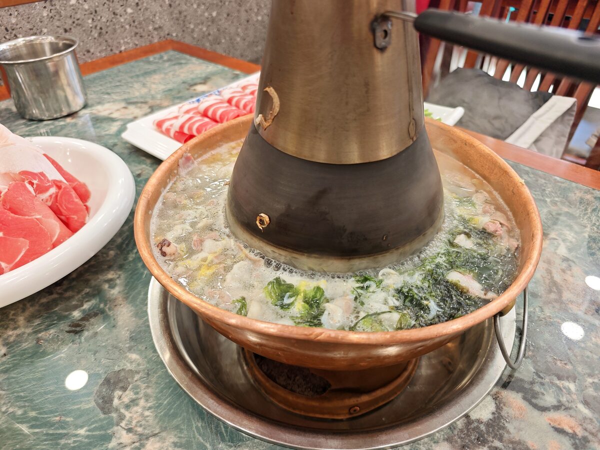 A northeastern-style copper hot pot with a central chimney, simmering broth and raw sliced meat beside it