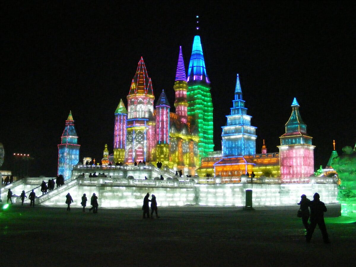 A tall ice castle with spires lit pink, blue and green at night in the Ice and Snow World, with visitors in silhouette