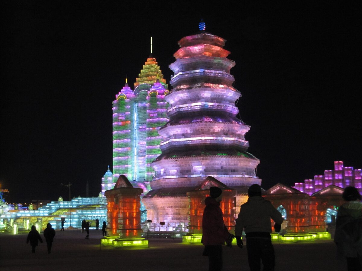 A tall ice pagoda and a green-lit ice tower glowing at night, with visitors in silhouette in front