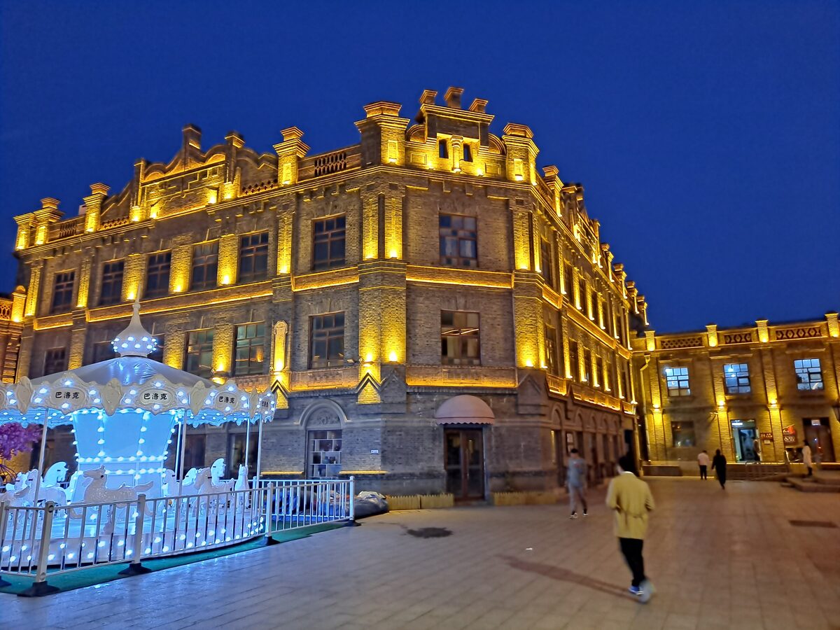 A corner building of the Chinese Baroque district floodlit in gold at dusk, with a small carousel in front
