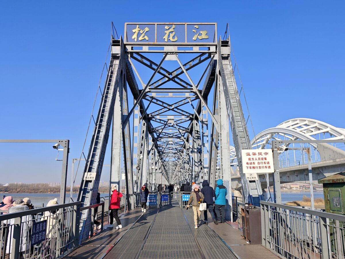 Walkers on the old Songhua River Railway Bridge, its steel truss entrance gate marked with the river's name in Chinese characters