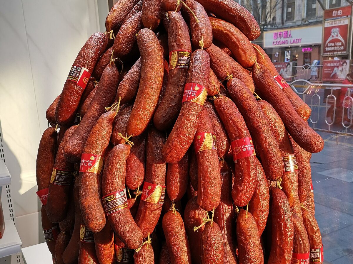 A tall bunch of dark red smoked sausages with red paper bands hanging in a shop window