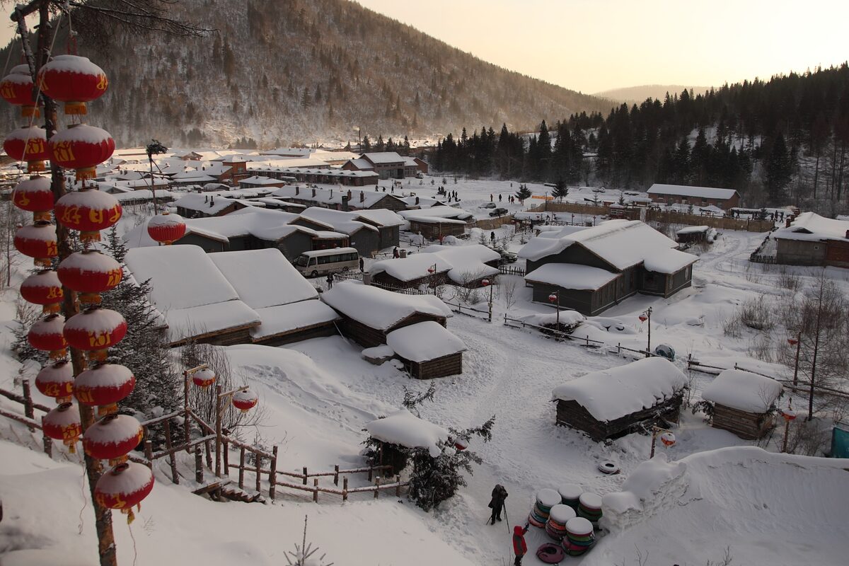 Snow-covered village houses on a forested hillside with red lanterns strung along a pole in the foreground