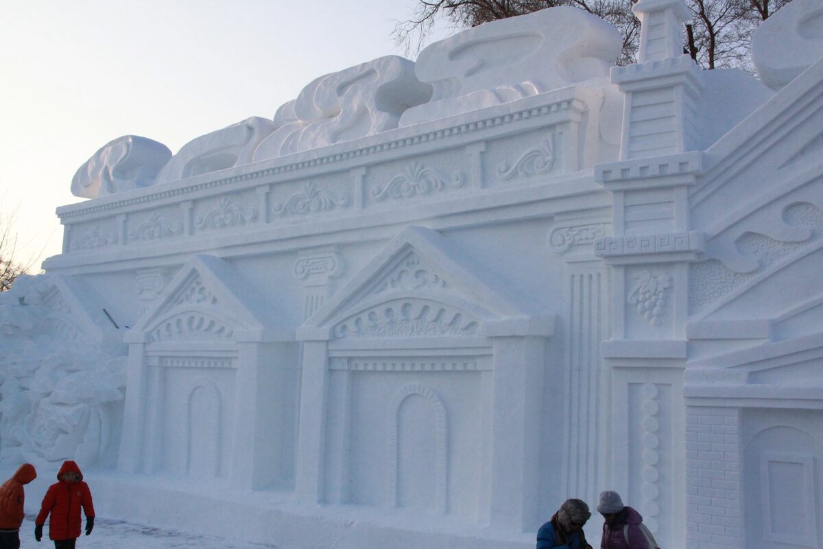 A huge snow sculpture carved as a columned building facade, with visitors at its foot
