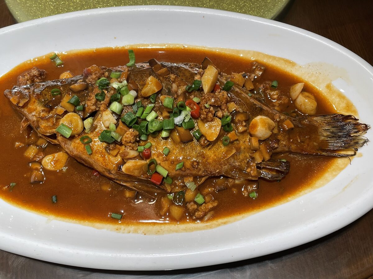 A whole braised mandarin fish on an oval plate, covered in a brown sauce with minced pork and scallions