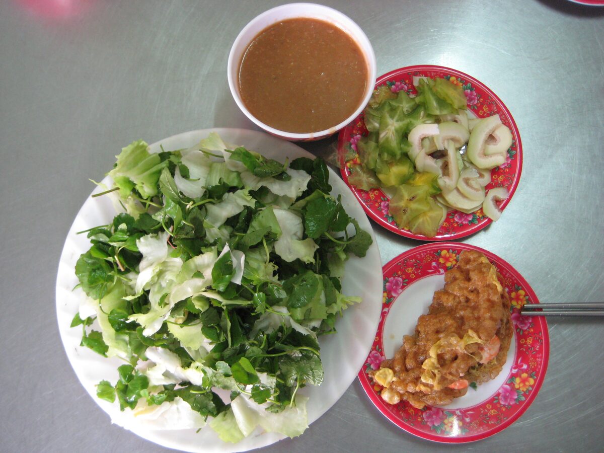 A banh khoai pancake under peanut sauce on a small plate, beside a big plate of fresh herbs and a dish of pickles