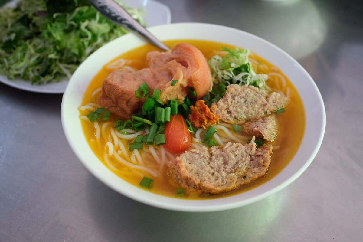 A bowl of bun bo Hue in orange-red broth with noodles, pork cake slices and herbs