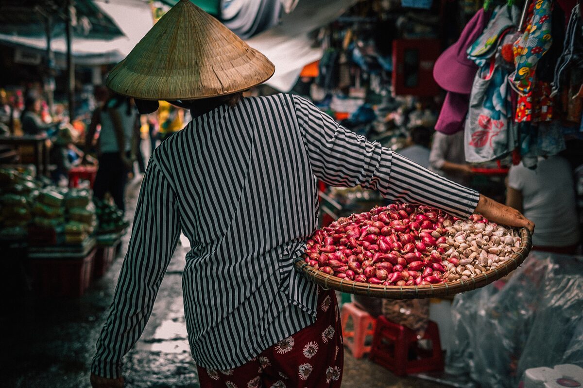 A vendor in a conical hat carrying a tray of red shallots and garlic through the stalls of Dong Ba Market