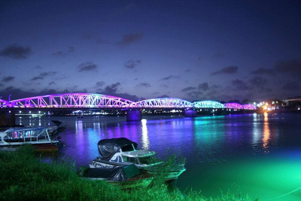Trang Tien Bridge lit in purple and blue across the Perfume River at dusk, boats moored on the bank