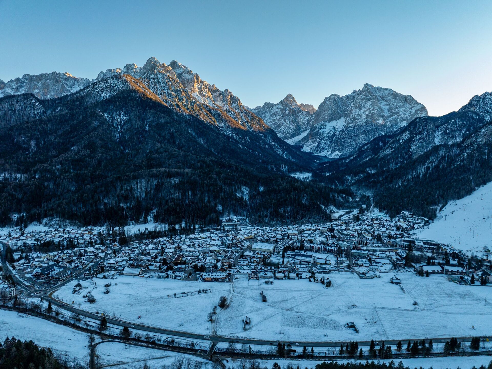 Aerial view of Kranjska Gora under snow on a clear winter day, with the village in the valley and the Julian Alps rising behind it