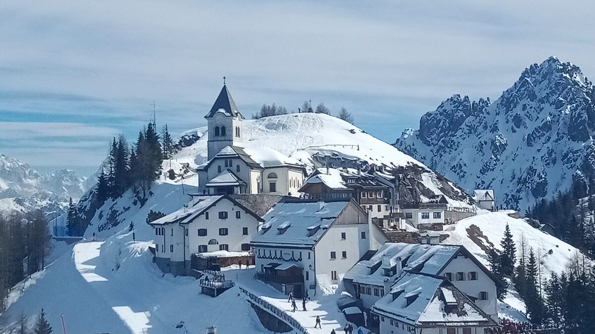 The Monte Lussari sanctuary and its village on a snowy hilltop above Tarvisio, with rocky peaks behind