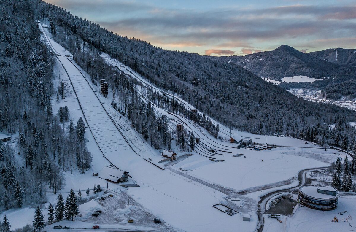 Aerial view of the Planica jumping hill under snow, the long takeoff ramp running down through the spruce forest