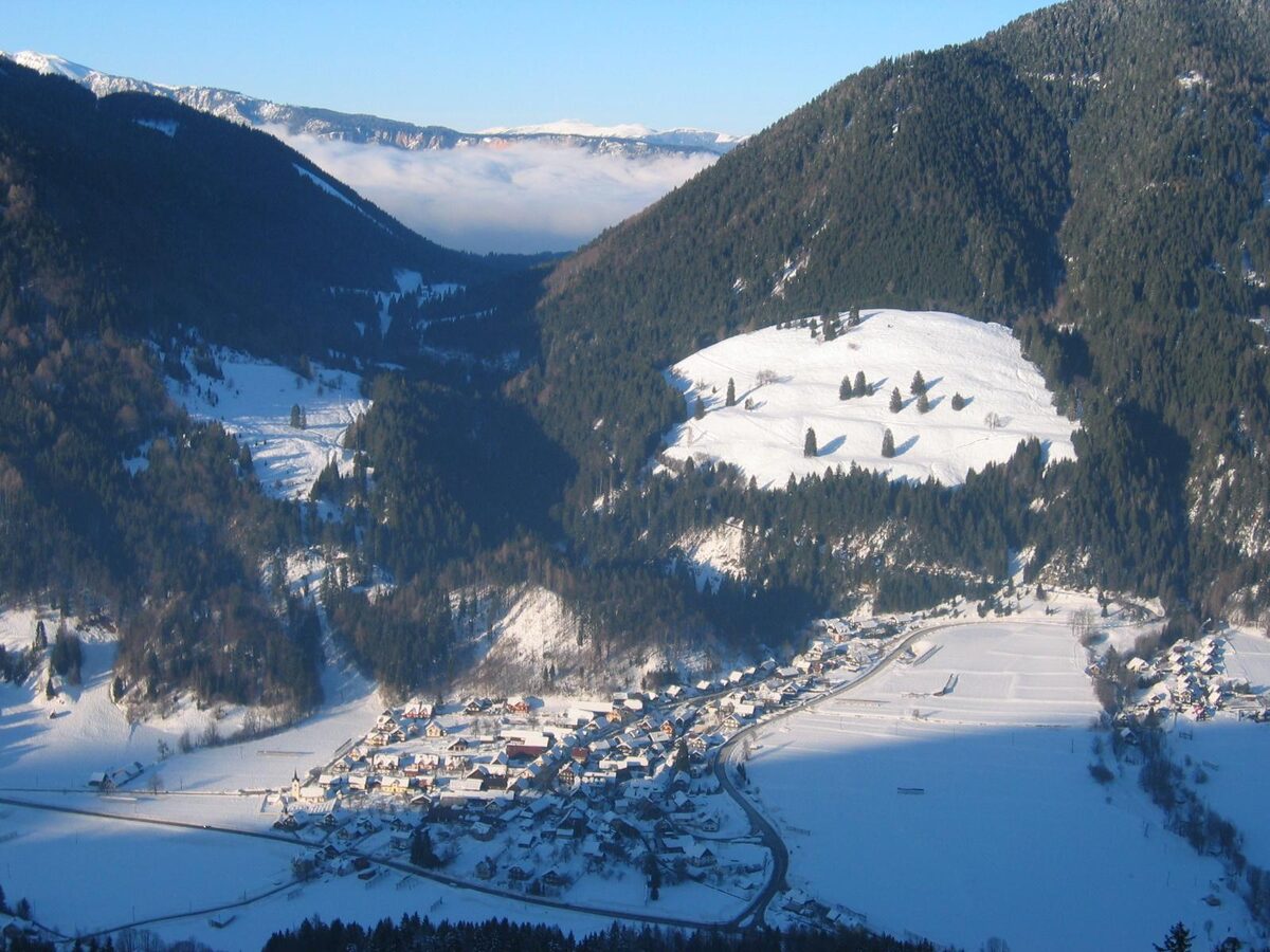 Podkoren village in a snowy valley seen from the Podkoren ski slope, with forested slopes and a snow-covered meadow above it