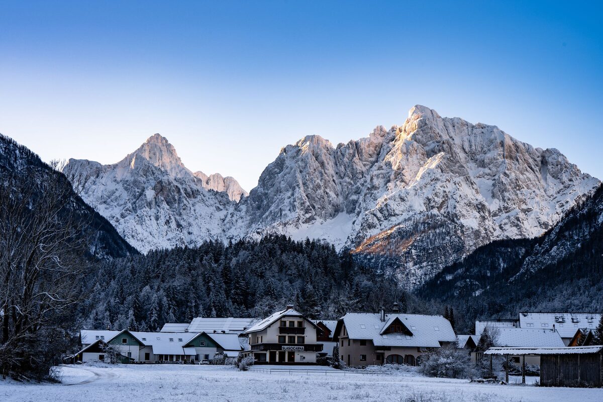 Snow-covered houses and fields below the snowy rock walls of the Julian Alps in low winter light