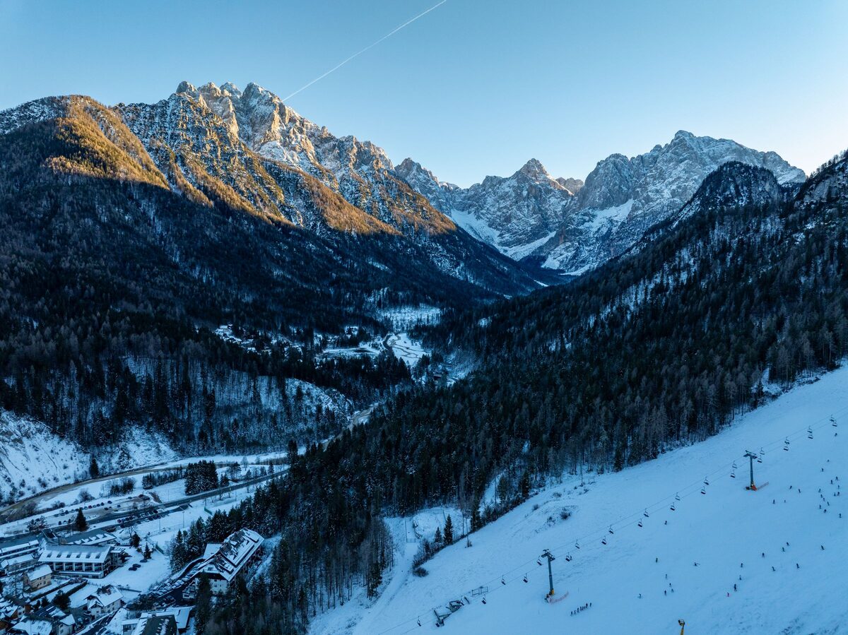 A snowy ski slope with a chairlift and skiers beside a forested valley, with sunlit Julian Alps peaks ahead