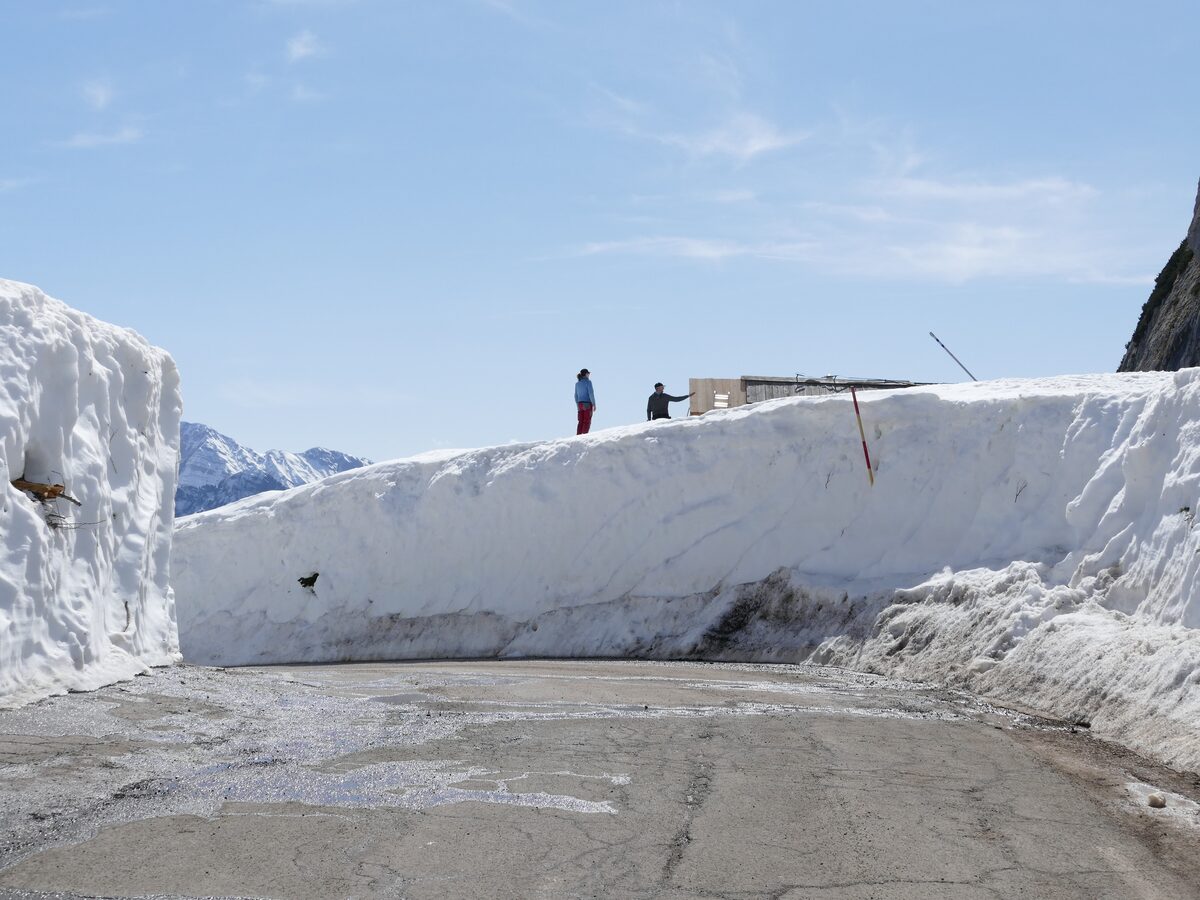 Snow walls lining the Vršič road, with two people standing on top of the snow bank under a blue sky