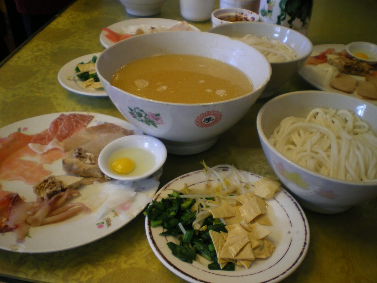 A bowl of broth beside rice noodles and plates of raw meat, egg and greens