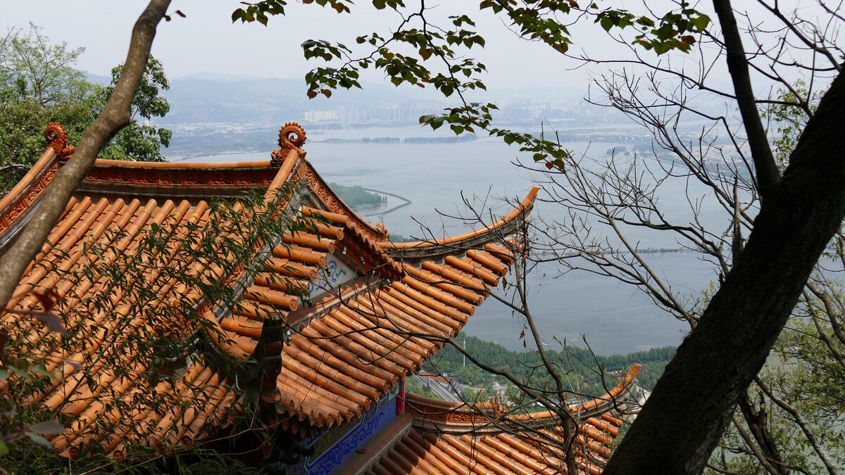 Orange tiled roofs of a Dragon Gate temple on the cliff, with Dianchi lake beyond the trees