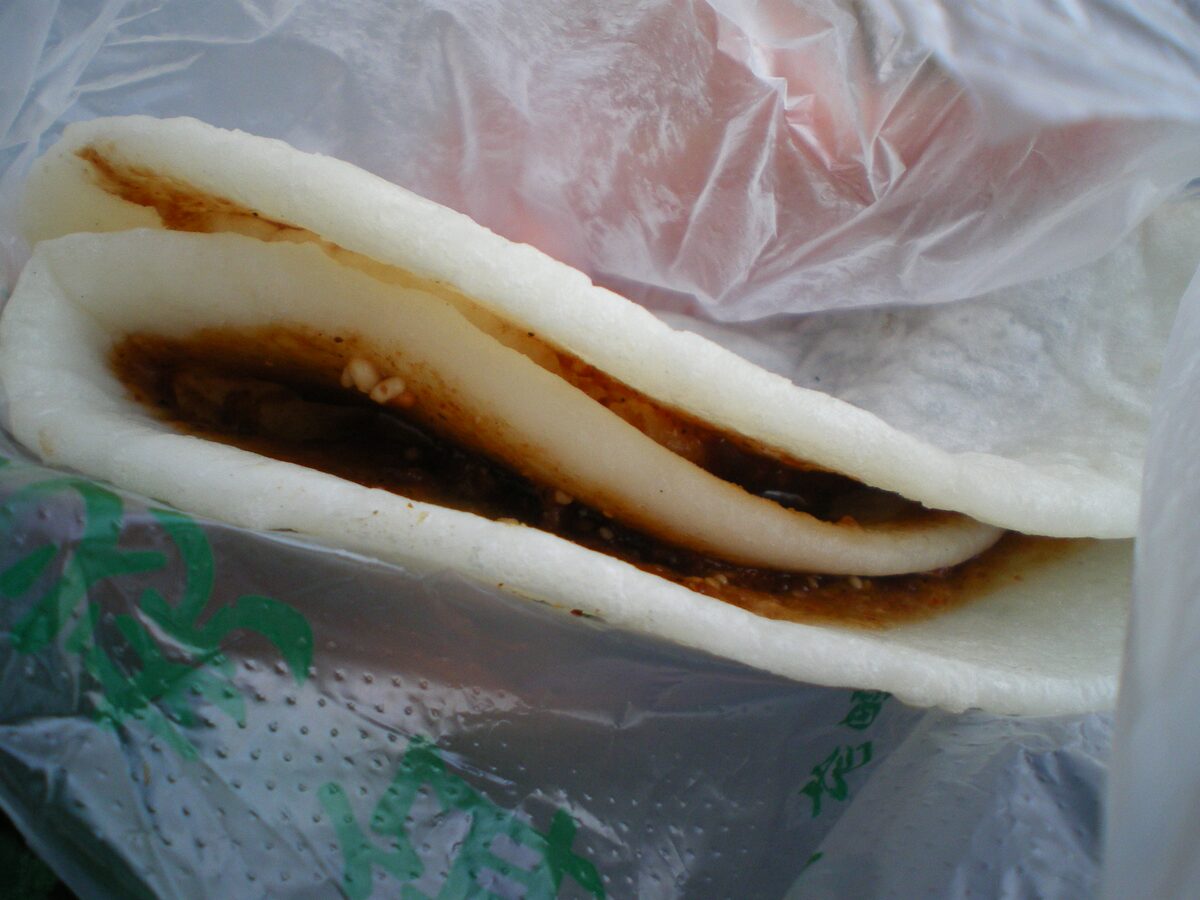 A grilled er kuai rice cake folded around dark sweet-spicy sauce, in a plastic bag
