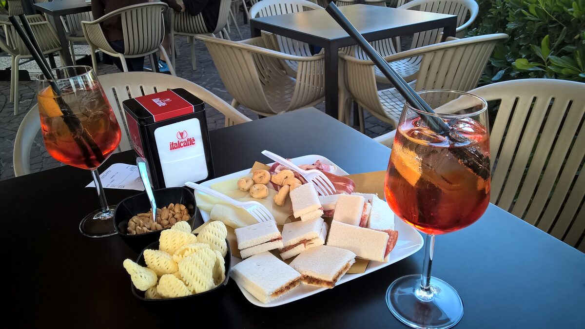 Two Aperol spritz glasses on a table with crisps and sandwiches