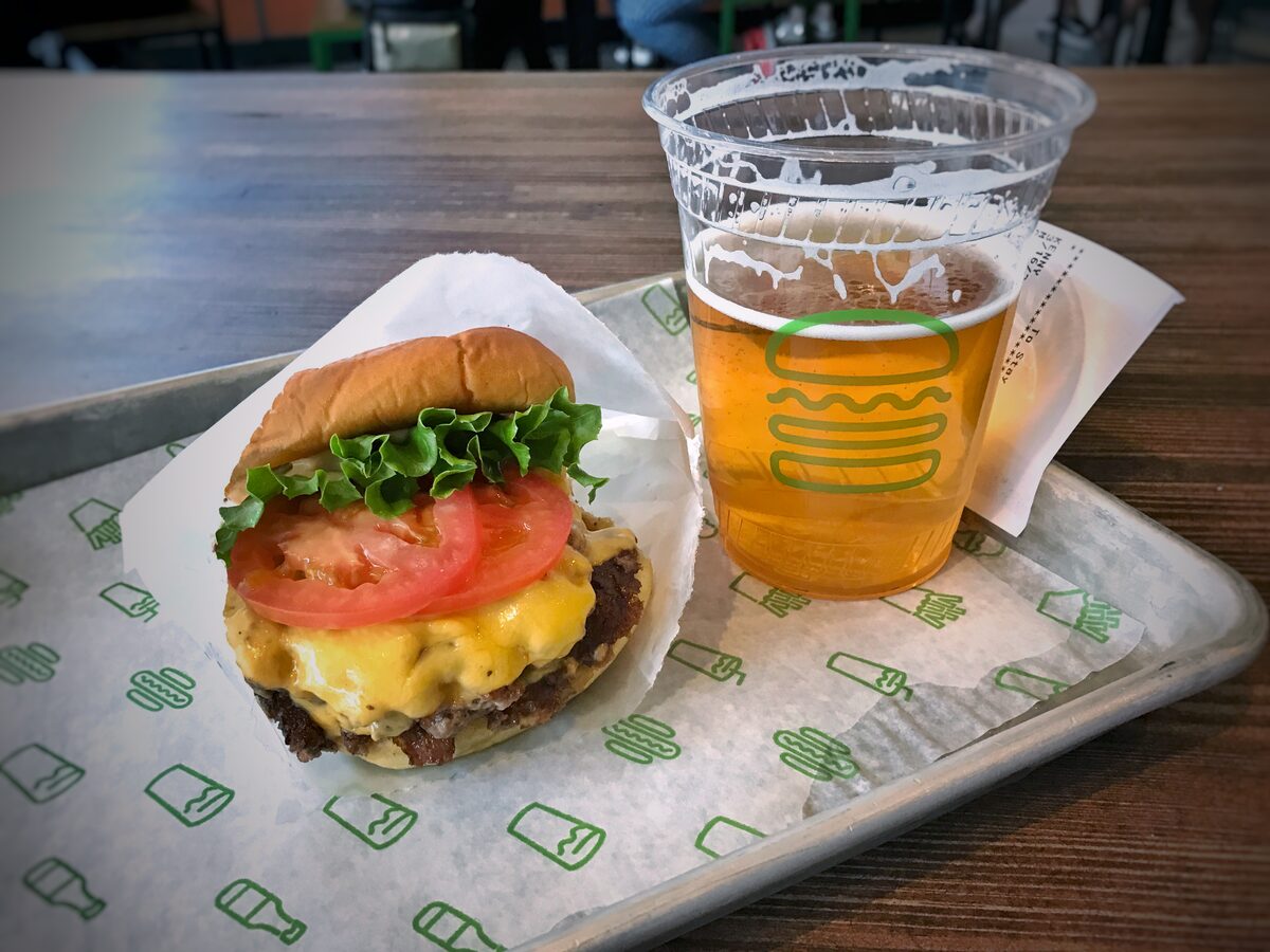 A cheeseburger with lettuce and tomato on a soft bun in paper, next to a glass of beer on a tray