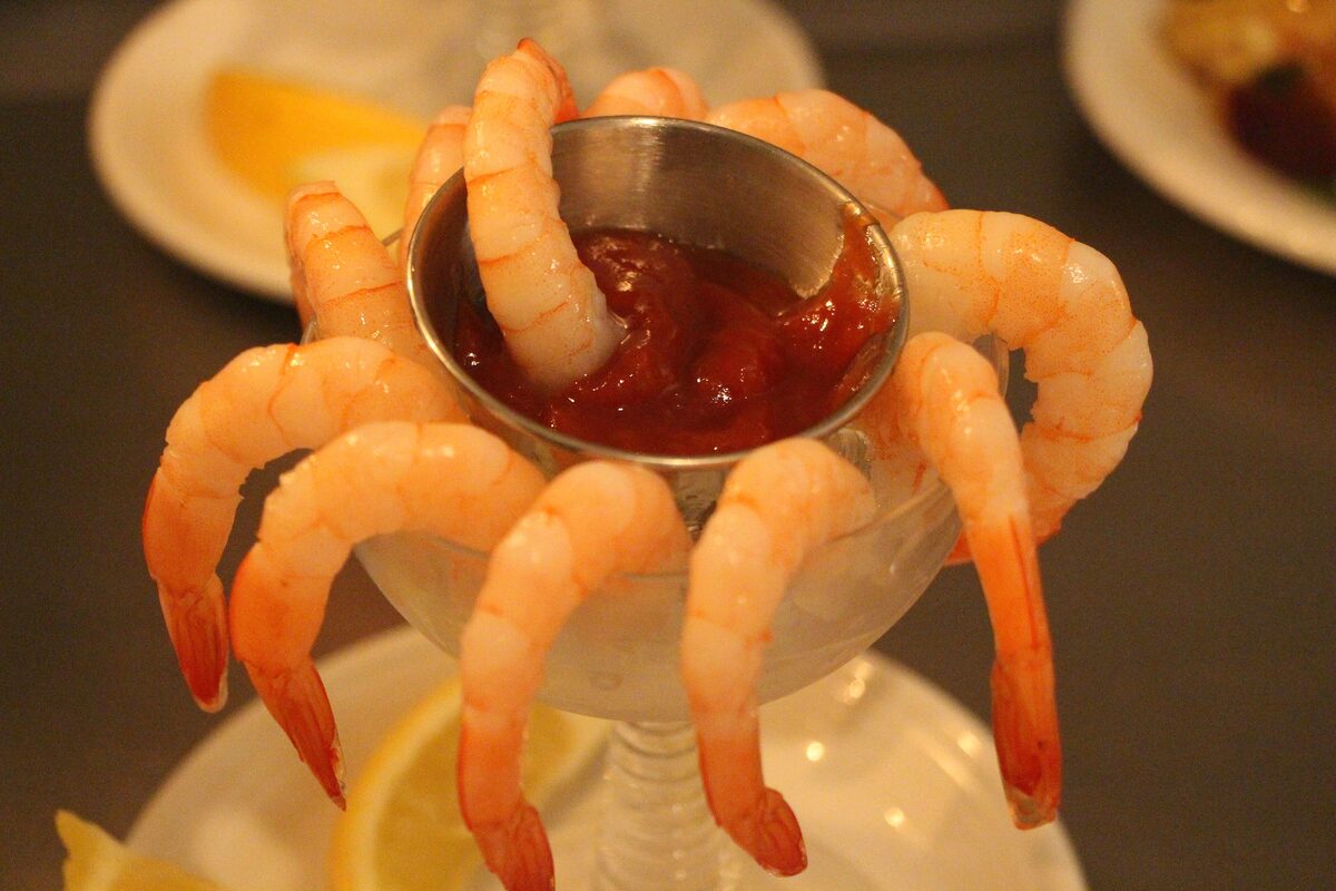 Shrimp hanging on the rim of a cocktail glass around a small cup of red cocktail sauce